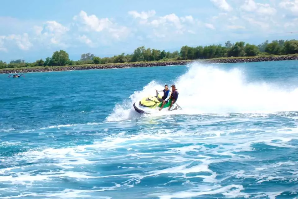 Two adventurers riding a jet ski at high speed across the clear blue waters of Bali, creating splashes under a sunny sky.